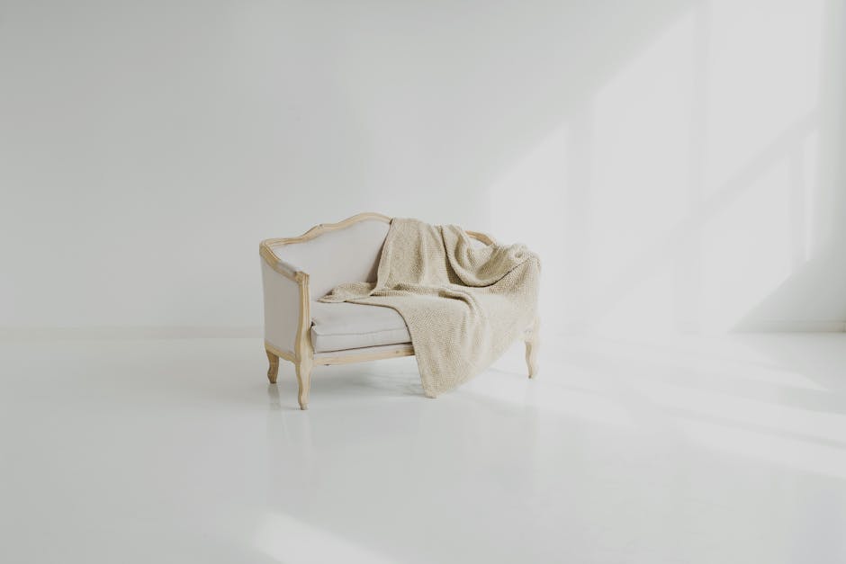 A vintage-style two-seater sofa with a cream-colored wooden frame and light upholstery positioned against a white wall in a bright, minimalistic room. A beige textured throw blanket is casually draped