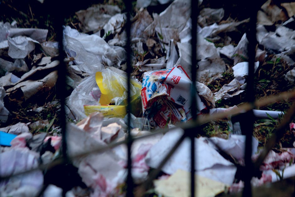 A close-up of a gated outdoor area with black metal bars, showing a ground cluttered with crumpled and discarded waste, including paper and plastic packaging, amidst fallen leaves. The waste appears u